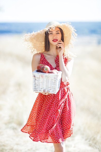 Retrato de uma jovem mulher bonita usando chapéu de palha no verão. Fêmea com maçãs frescas na natureza.