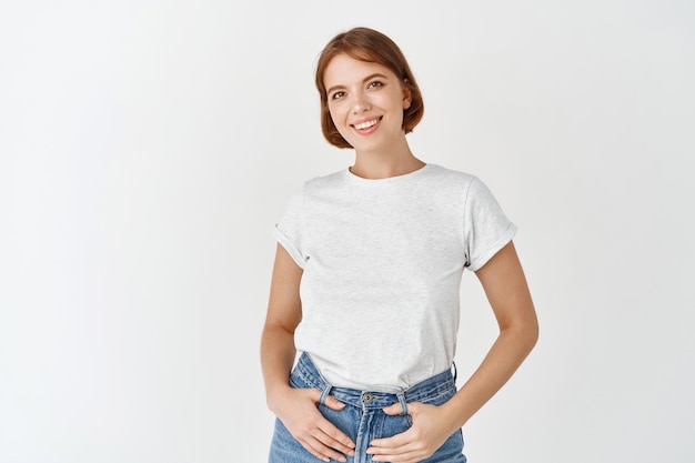 Retrato de uma jovem mulher cândida em uma camiseta, segurando as mãos em jeans e sorrindo feliz, em pé na parede branca