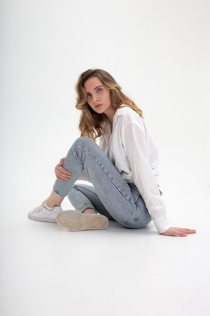 Retrato de uma jovem mulher caucasiana pensativa, posando de camisa e calça jeans, sentada no chão branco. testes de modelo de garota bonita com roupas básicas no ciclorama