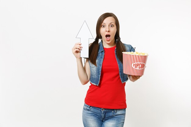 Retrato de uma jovem mulher chocada em roupas casuais, assistindo a um filme, segurando um balde de pipoca, apontando a seta branca para cima em copyspace isolado no fundo branco. Emoções no conceito de cinema.