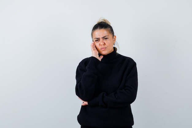 Retrato de uma jovem mulher com as mãos na bochecha, com uma blusa de gola alta e olhando pensativa para a frente