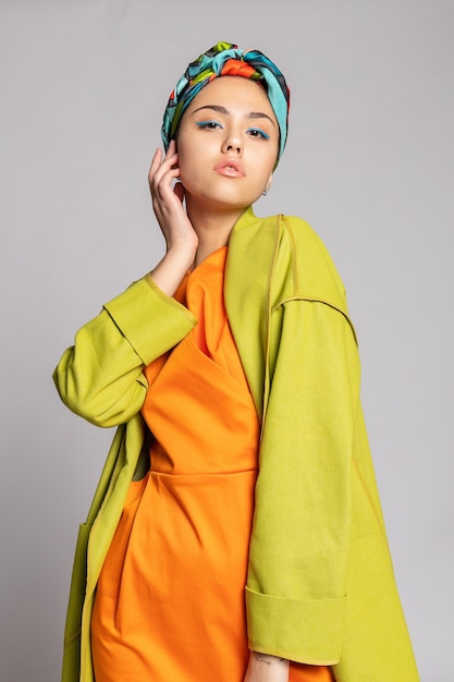 Retrato de uma jovem mulher com maquiagem brilhante e um lenço na moda. Fundo claro. Beleza, moda, conceito de maquiagem. garota com um casaco verde brilhante, vestido laranja brilhante.