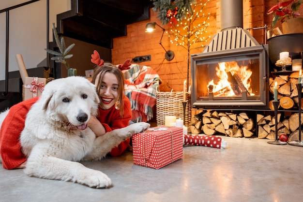 Retrato de uma jovem mulher com seu lindo cachorro branco desempacotando caixas de presente perto da lareira durante um feliz ano novo em casa