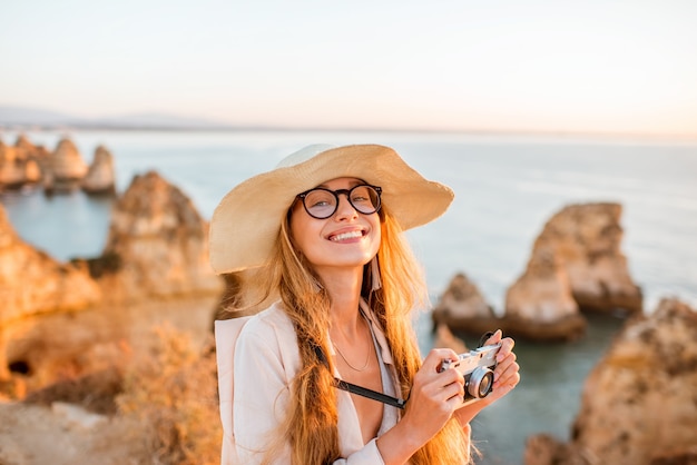 Retrato de uma jovem mulher com uma câmera fotográfica, desfrutando de uma bela vista da costa rochosa durante o nascer do sol em Lagos, no sul de Portugal