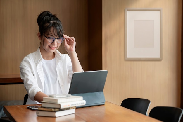 Retrato de uma jovem mulher de negócios com a mão tocar seus óculos e trabalhar no computador enquanto está sentado em sua mesa de escritório.