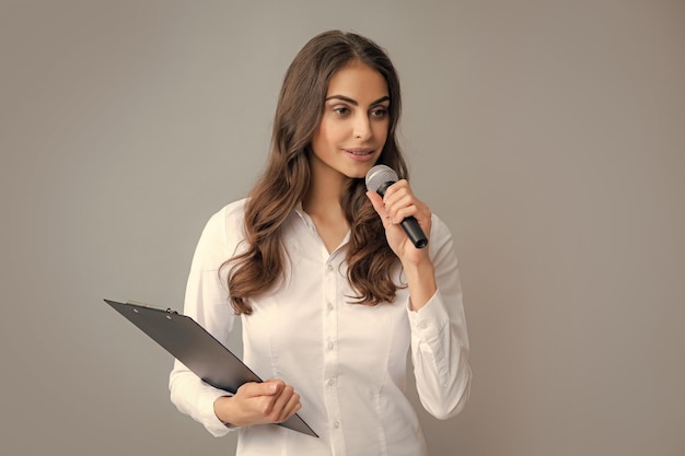 Retrato de uma jovem mulher de negócios sorridente fazendo apresentação com prancheta e microfone isolado de fundo cinza