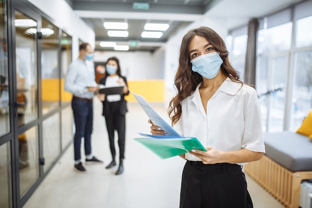 Retrato de uma jovem mulher de negócios usando uma máscara médica, verificando papéis, tendências e perspectivas de negócios em um corredor de escritório.