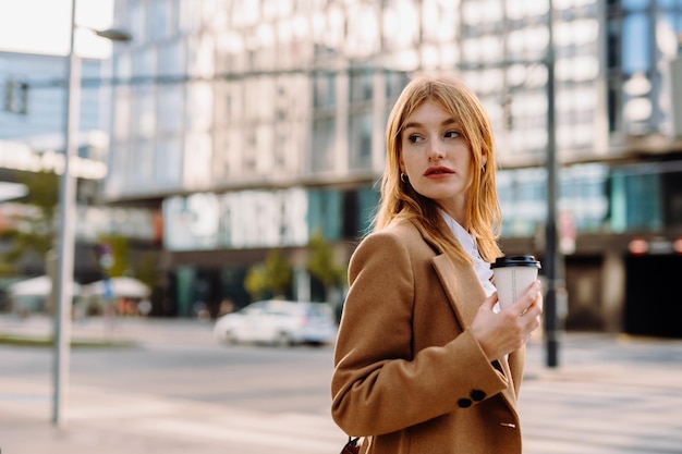 Retrato de uma jovem mulher de negócios vestindo um casaco de cor de camelo andando na rua de uma cidade europeia