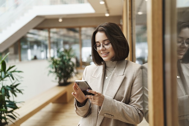 Retrato de uma jovem mulher de sucesso trabalhando para a empresa, esperando o colega de trabalho para o almoço no corredor, verificando mensagens no celular, usando o smartphone e sorrindo satisfeito.