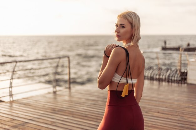 Retrato de uma jovem mulher esportiva em roupas esportivas com uma corda de pular. Esportista posando na praia ao nascer do sol