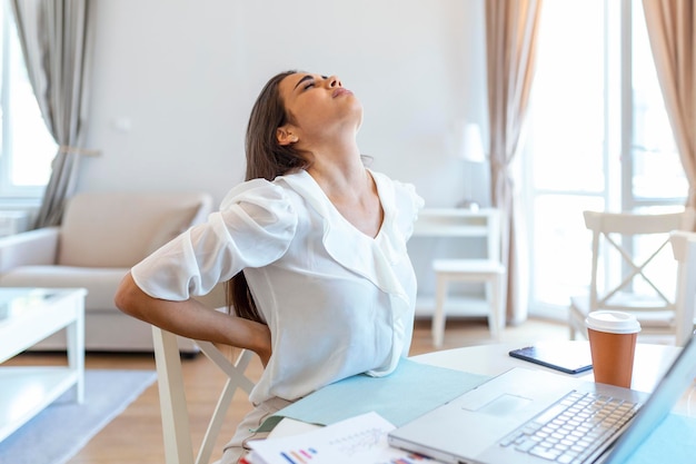 Retrato de uma jovem mulher estressada sentada na mesa do escritório em casa em frente ao laptop, tocando as costas doloridas com expressão de dor, sofrendo nas costas, dor na coluna depois de trabalhar no pc