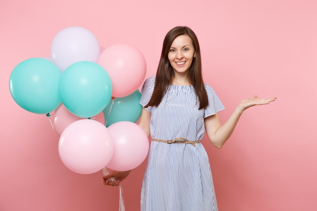 Retrato de uma jovem mulher fascinante feliz em um vestido azul segurando balões de ar coloridos, apontando a mão de lado no espaço da cópia, isolado em um fundo rosa tendência brilhante. conceito de festa de aniversário.