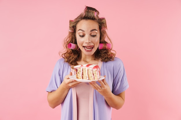 Retrato de uma jovem mulher feliz em bobes sorrindo e segurando um bolo isolado no rosa