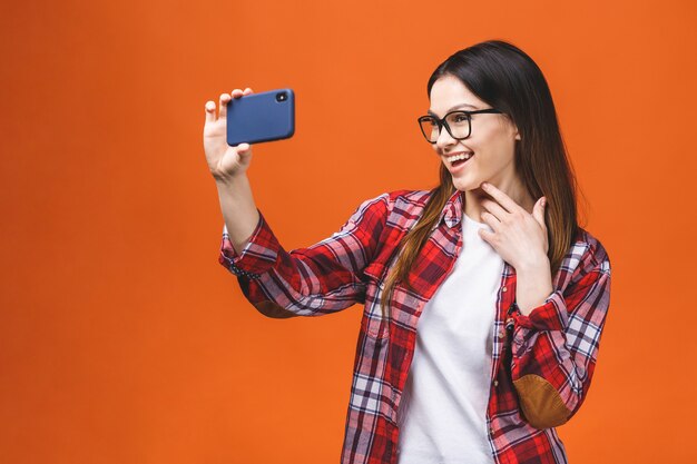 Retrato de uma jovem mulher feliz fazendo selfie no smartphone, isolado contra um fundo laranja.