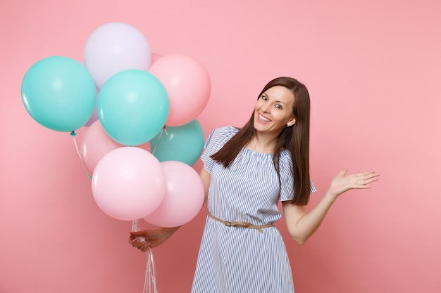 Retrato de uma jovem mulher feliz sorridente, usando um vestido azul, segurando balões de ar coloridos, apontando a mão de lado no espaço da cópia, isolado no fundo rosa tendência brilhante. Conceito de festa de aniversário.