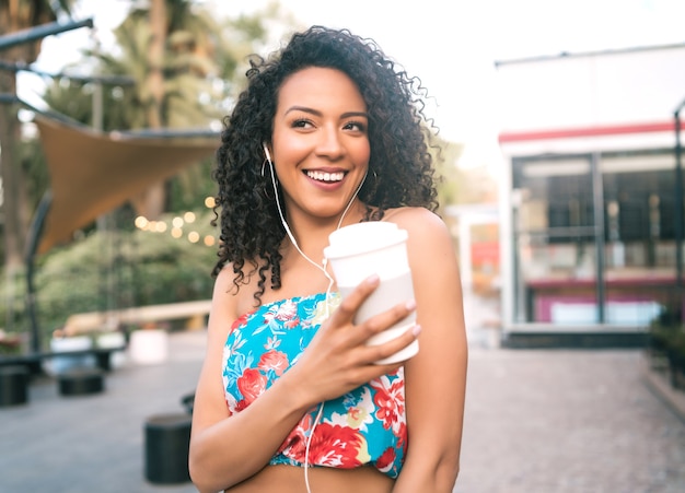 Retrato de uma jovem mulher latino afro-americana, ouvindo música com fones de ouvido, segurando uma xícara de café ao ar livre na rua.