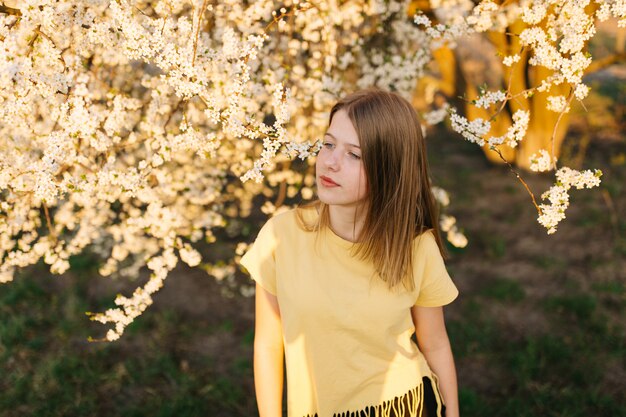 Retrato de uma jovem mulher loira bonita perto de árvore florescendo com flores brancas em um dia ensolarado.