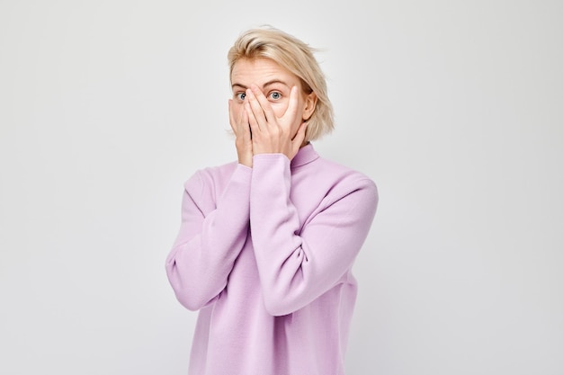 Foto retrato de uma jovem mulher loira cobrindo os olhos com a mão espreitando através dos dedos isolados no fundo branco do estúdio