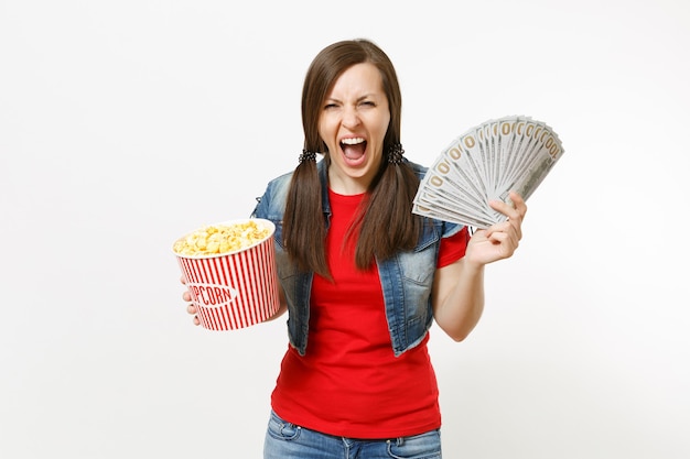 Retrato de uma jovem mulher louca em roupas casuais, assistindo a um filme de filme, segurando um balde de pipoca e um pacote de dólares, dinheiro vivo, gritando, isolado no fundo branco. Emoções no conceito de cinema.