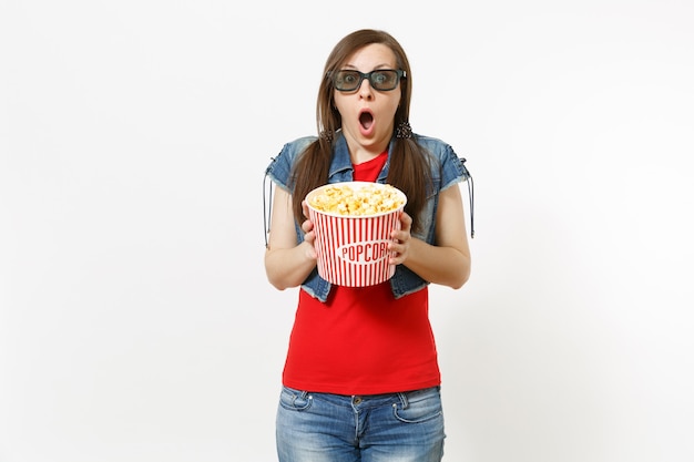Retrato de uma jovem mulher morena atraente chocada em óculos 3d imax e roupas casuais, assistindo a um filme de cinema, segurando um balde de pipoca isolado no fundo branco. Emoções no conceito de cinema.