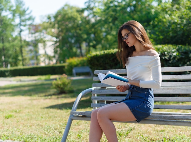 Retrato de uma jovem mulher morena atraente lendo livro, sentado no parque verde de verão.