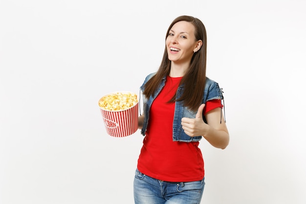 Retrato de uma jovem mulher morena atraente sorridente em roupas casuais, assistindo a um filme de cinema, segurando um balde de pipoca e aparecendo o polegar isolado no fundo branco. Emoções no conceito de cinema.