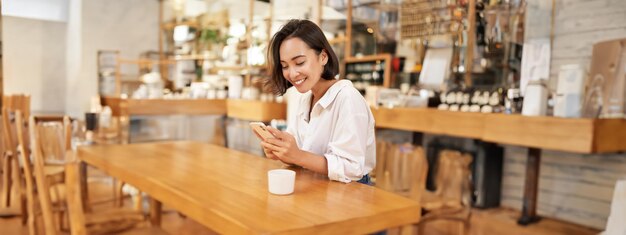 Retrato de uma jovem mulher morena sentada com café e usando um smartphone em um café conversando