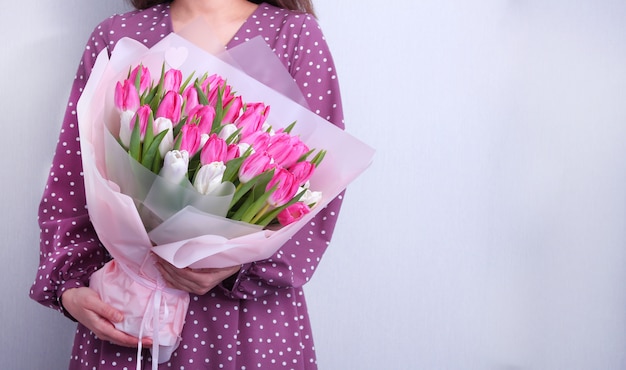 Retrato de uma jovem mulher no vestido roxo que guarda o ramalhete de tulipas cor-de-rosa e brancas no cinza. Cartão da mola. Páscoa, conceito de flor de primavera. Dia das mães ou da mulher.