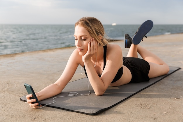 Retrato de uma jovem mulher ouvindo música no celular, deitada na esteira enquanto se exercita no cais, perto da beira-mar, pela manhã