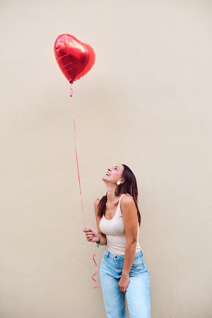 Retrato de uma jovem mulher segurando um balão em forma de coração em pé ao ar livre contra uma parede. Conceito de amor.