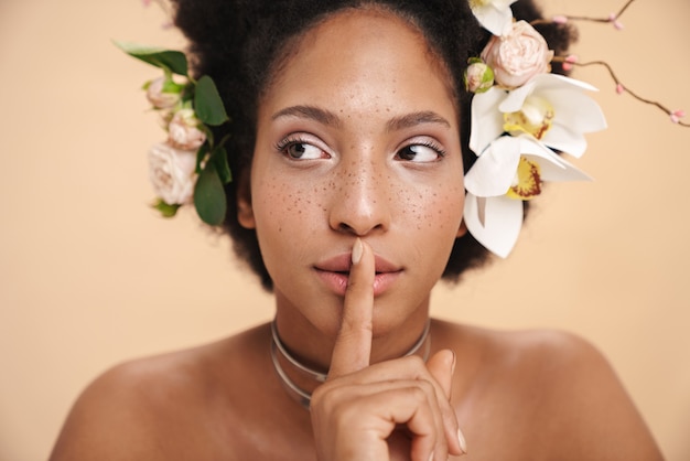 Retrato de uma jovem mulher seminua com flores no cabelo fazendo um gesto silencioso com o dedo nos lábios