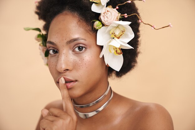 Retrato de uma jovem mulher seminua com flores no cabelo fazendo um gesto silencioso com o dedo nos lábios