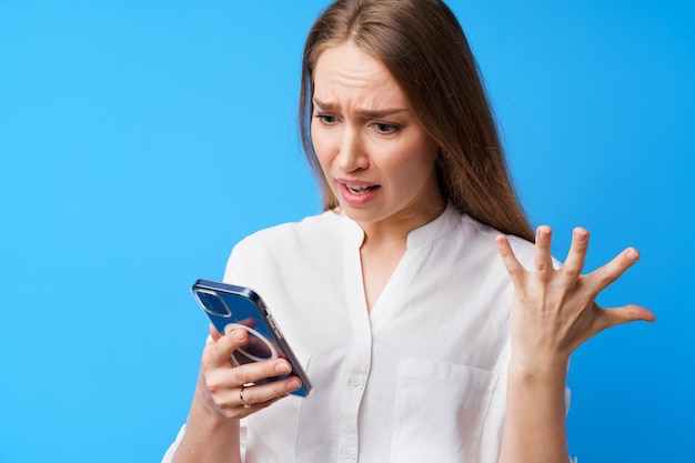 Retrato de uma jovem mulher zangada usando o telefone irritada enviando mensagens de texto com alguém com fundo azul