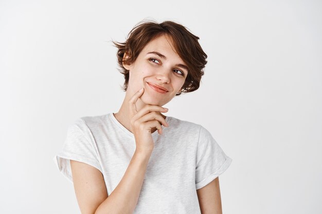 Retrato de uma jovem natural com penteado curto, parecendo pensativo à parte, sorrindo e sonhando com algo, parado pensativo na parede branca