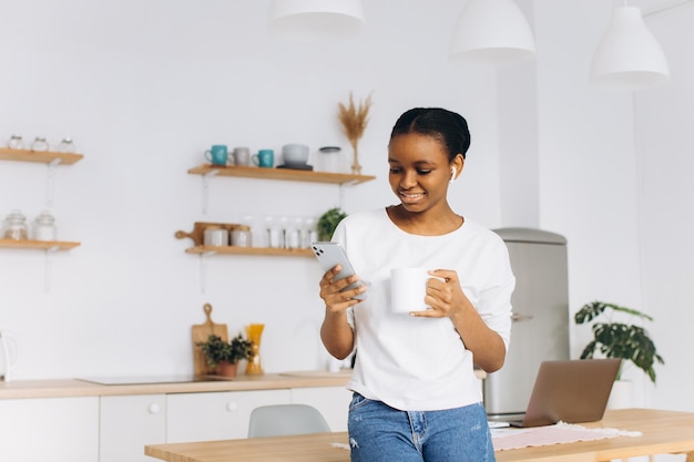 Retrato de uma jovem negra alegre segurando um telefone e tomando café na cozinha