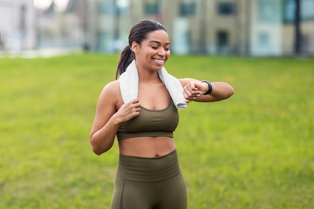 Retrato de uma jovem negra desportiva com toalha verificando seu smartwatch ou rastreador de fitness no parque da cidade