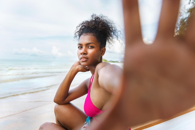 Foto retrato de uma jovem negra relaxando em uma cadeira de madeira na praia enquanto olha para a câmera