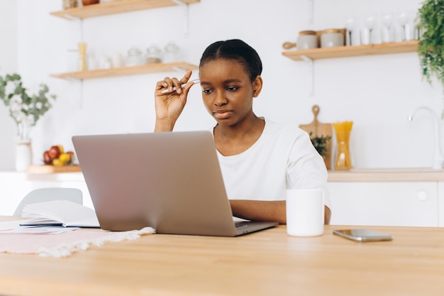 Retrato de uma jovem negra sentada na cozinha e trabalhando em um laptop em casa.