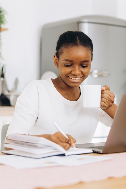 Retrato de uma jovem negra sentada na cozinha escreve algo e trabalhando em um laptop em casa.