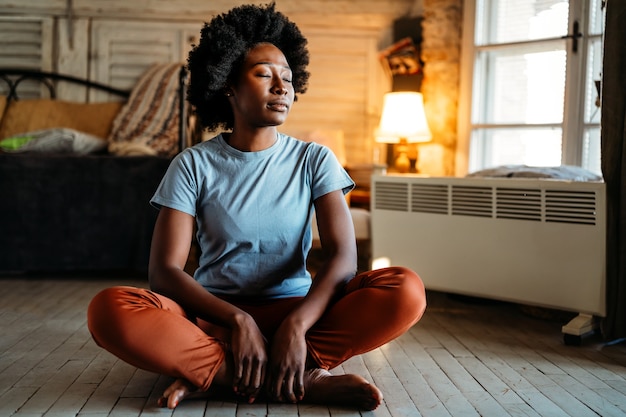 Foto retrato de uma jovem negra sorridente, sentada no chão, praticando ioga em casa