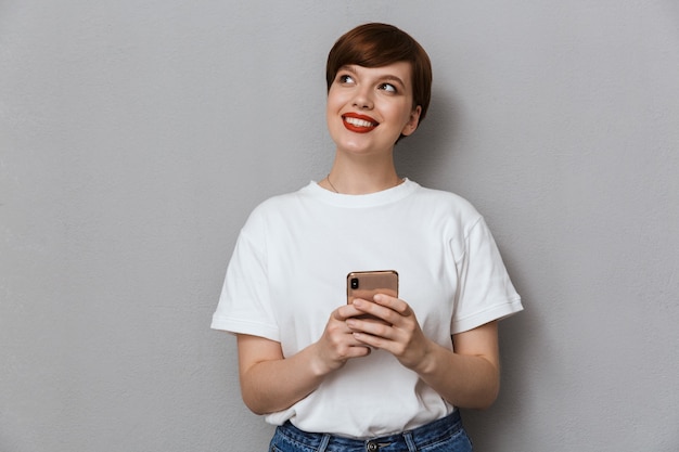 Retrato de uma jovem pensativa sorrindo e segurando o celular isolado sobre uma parede cinza