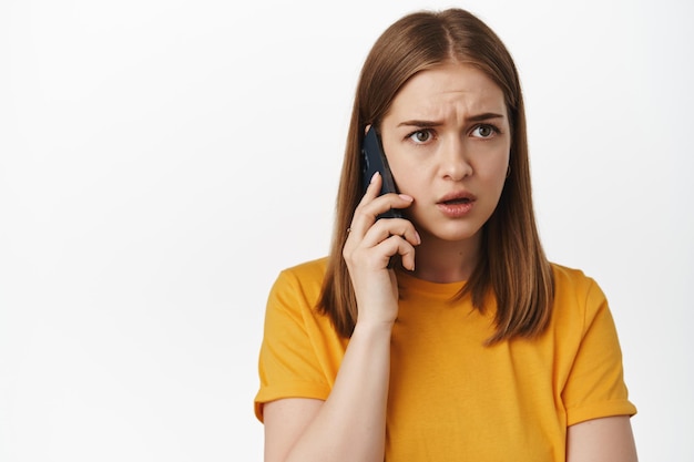 Retrato de uma jovem preocupada ouvindo com cara preocupada, receber um telefonema alarmante, falando no smartphone, vestindo camiseta amarela, de pé sobre fundo branco