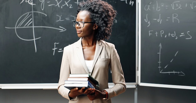 Foto retrato de uma jovem professora afro-americana de óculos, olhando para a câmera na sala de aula e segurando livros didáticos. quadro-negro com fórmulas no fundo. conceito de escolaridade. livros nas mãos da mulher.