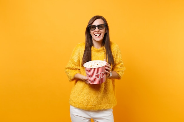 Retrato de uma jovem rindo em óculos 3d imax, assistindo a um filme, comer pipoca de balde isolado em fundo amarelo brilhante. Emoções sinceras de pessoas no cinema, conceito de estilo de vida. Copie o espaço.