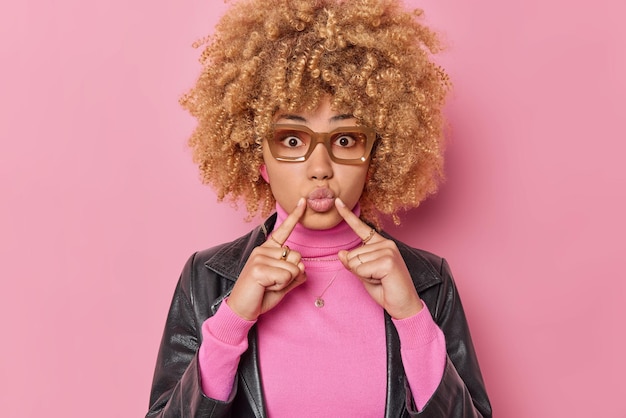 Foto retrato de uma jovem romântica de cabelos encaracolados linda aponta o dedo indicador para lábios arredondados usa óculos de gola alta jaqueta de couro isolada sobre fundo rosa. conceito de expressões de rosto humano