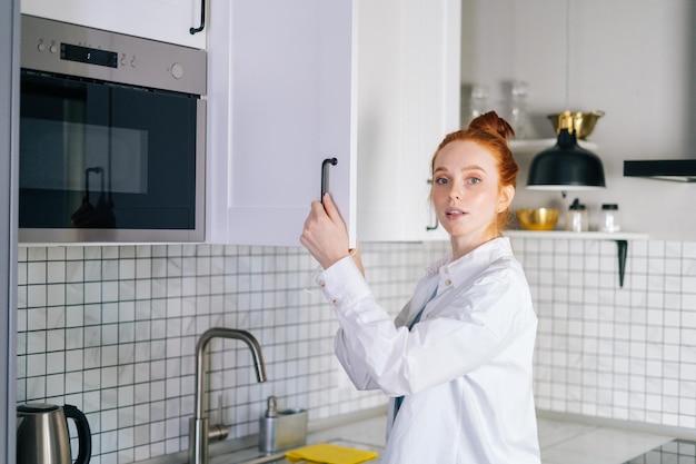 Retrato de uma jovem ruiva abrindo a porta do armário de cozinha na sala de cozinha leve