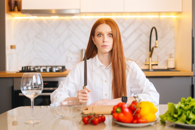 Retrato de uma jovem ruiva atraente com olhos malucos, segurando uma faca grande, sentado à mesa com