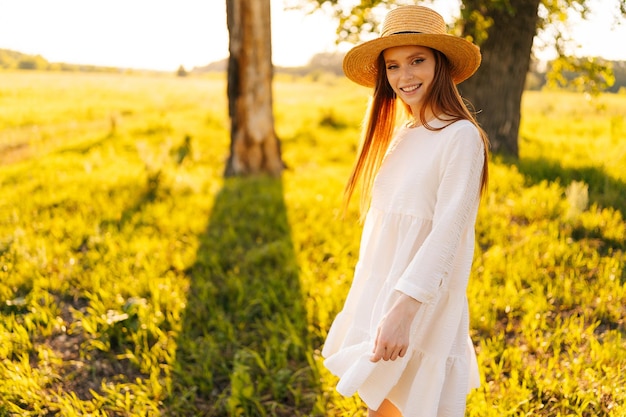 Retrato de uma jovem ruiva encantadora com chapéu de palha e vestido branco em pé posando em um belo prado de grama verde olhando para a câmera