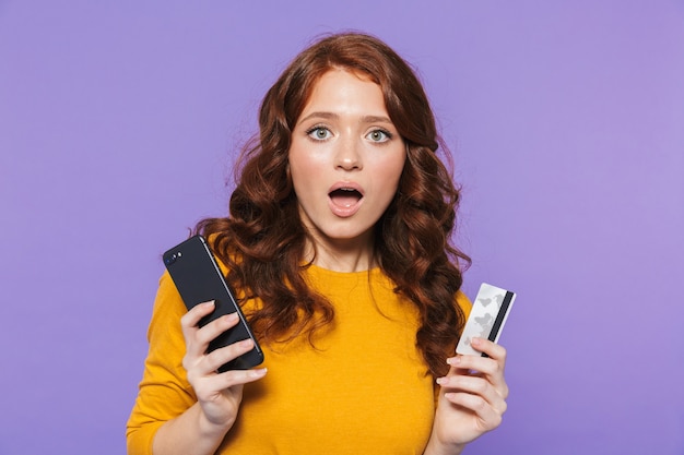 Retrato de uma jovem ruiva muito alegre em pé sobre a violeta, usando o celular, mostrando o cartão de crédito