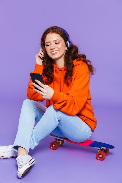 Retrato de uma jovem ruiva sentada no skate sobre a violeta, ouvindo música com fones de ouvido